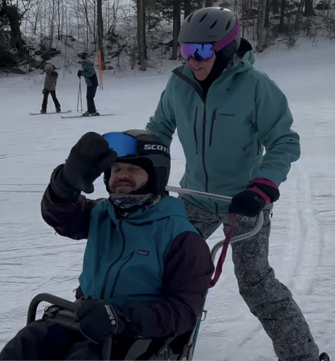 Travis waiving while on a bi-ski. Unknown person is guiding the sled.